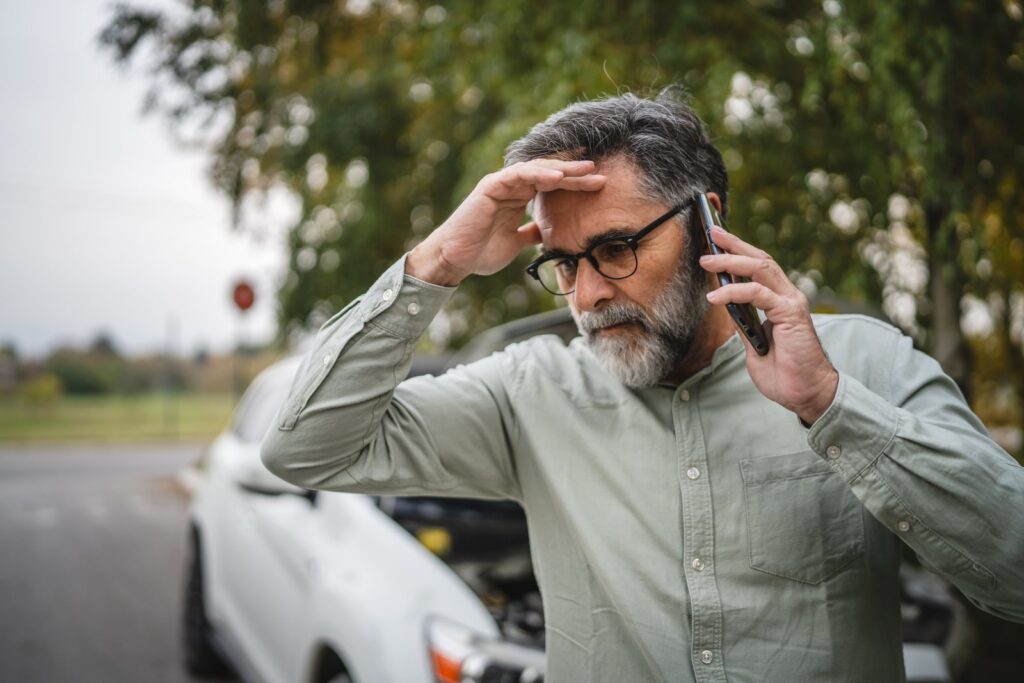 Driver with a hand on his forehead calling for help on his phone beside a damaged car after a crash